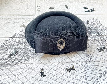 May include: A black hat with a black veil and a silver bat brooch. The hat is made of a soft, felt-like material and has a wide brim.