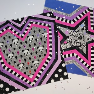 May include: Two fabric squares with a heart and a star design. The heart is pink, black, and white with a panda print. The star is pink, black, and white with a floral print. Both squares have a purple, black, and white striped border.