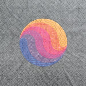May include: A circular design featuring layered, curved segments in shades of blue, purple, pink, and orange, with intricate patterns. The design is set against a gray, textured background.