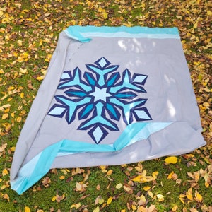 May include: A gray quilt with a large snowflake design in shades of blue. The snowflake is centered on the quilt, which has a light blue border. The quilt is displayed on a bed of green grass and fallen leaves.