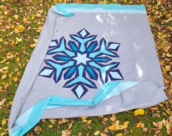 May include: A gray quilt with a large snowflake design in shades of blue. The snowflake is centered on the quilt, which has a light blue border. The quilt is displayed on a bed of green grass and fallen leaves.