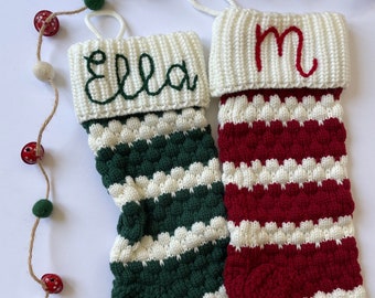 Op de afbeelding: Twee gebreide kerstsokken met een textuur van noppensteken. De ene sok is donkergroen en wit met de naam Ella in het groen geborduurd. De andere is rood en wit met de letter M in het rood geborduurd. Beide hebben witte boorden aan de bovenkant.