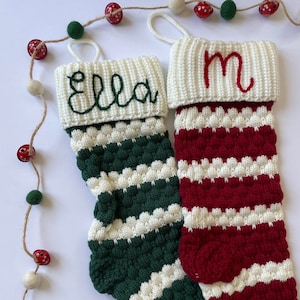 May include: Two knitted Christmas stockings featuring a textured bobble stitch design. One stocking is dark green and white with the name Ella embroidered in green. The other is red and white with the letter M embroidered in red. Both have white cuffs.