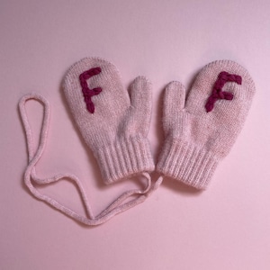 May include: A pair of pale pink knitted mittens, each with a dark pink embroidered letter "F". The mittens are joined by a matching pink cord. The background is a solid, light pink colour.