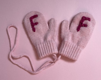 May include: A pair of pale pink knitted mittens, each with a dark pink embroidered letter "F". The mittens are joined by a matching pink cord. The background is a solid, light pink colour.