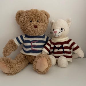 May include: A brown teddy bear wearing a blue and white striped sweater with the letter "E" embroidered on it, sitting next to a white cat wearing a red and white striped sweater with the letter "M" embroidered on it.