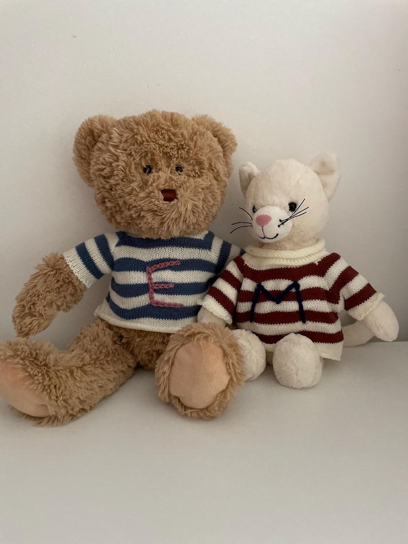 May include: A brown teddy bear wearing a blue and white striped sweater with the letter "E" embroidered on it, sitting next to a white cat wearing a red and white striped sweater with the letter "M" embroidered on it.
