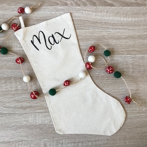 May include: A white canvas Christmas stocking with the name "Max" embroidered in black script. The stocking is decorated with a red and green garland.