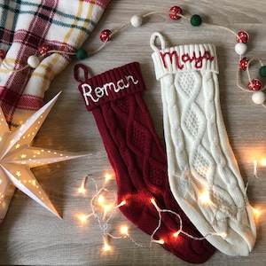 May include: Two cable-knit Christmas stockings, one burgundy and one cream, with the names "Roman" and "Maya" embroidered in white and red. A star-shaped paper lantern and string lights add to the festive scene. A plaid blanket and garland complete the holiday decor.