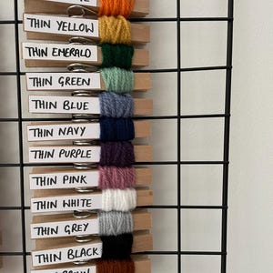May include: A wall organiser with wooden pegs holding different colours of yarn. The yarn colours are red, orange, yellow, green, blue, navy, purple, pink, white, grey, black, brown, and cream. The labels on the pegs read "Thin Red", "Thin Orange", "Thin Yellow", "Thin Emerald", "Thin Green", "Thin Blue", "Thin Navy", "Thin Purple", "Thin Pink", "Thin White", "Thin Grey", "Thin Black", "Thin Brown", and "Thin Cream".