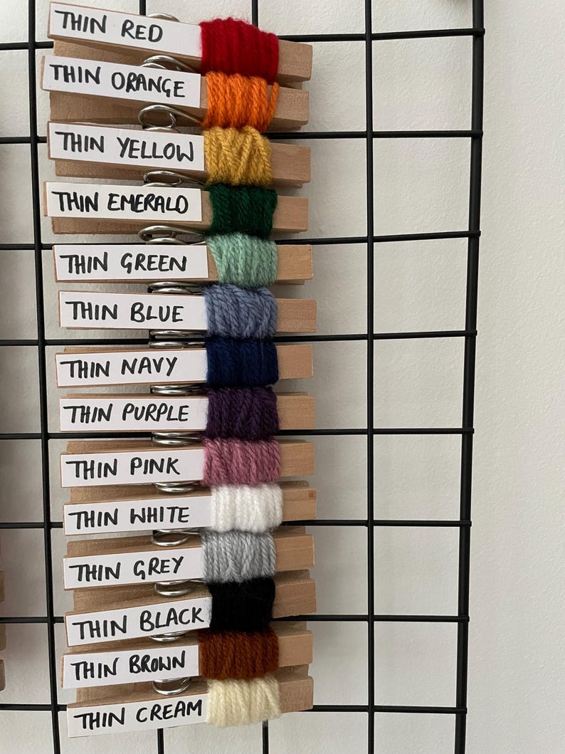 May include: A wall organiser with wooden pegs holding different colours of yarn. The yarn colours are red, orange, yellow, green, blue, navy, purple, pink, white, grey, black, brown, and cream. The labels on the pegs read "Thin Red", "Thin Orange", "Thin Yellow", "Thin Emerald", "Thin Green", "Thin Blue", "Thin Navy", "Thin Purple", "Thin Pink", "Thin White", "Thin Grey", "Thin Black", "Thin Brown", and "Thin Cream".