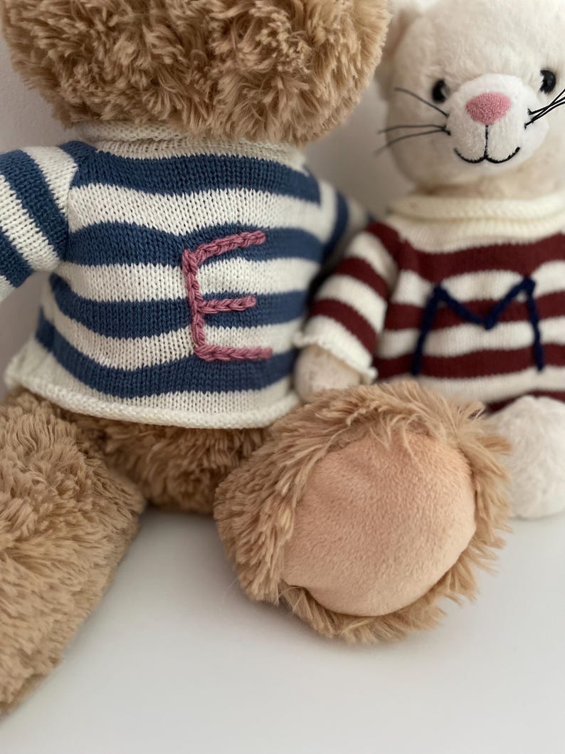 May include: Two plush toys, a brown bear wearing a blue and white striped sweater with a pink embroidered letter 'E' and a white cat wearing a red and white striped sweater with a blue embroidered letter 'M'.