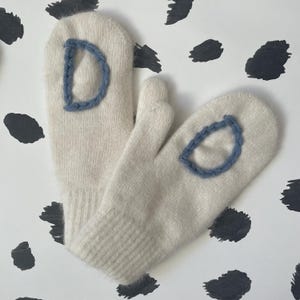 May include: A pair of white knitted mittens with blue embroidered letters "D" on each mitten.