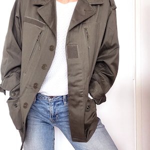 May include: An olive green utility jacket featuring a collared neckline, long sleeves, and a button-down front closure. The jacket includes two vertical zippered chest pockets, two lower flap pockets with snap buttons, and rectangular velcro patches.