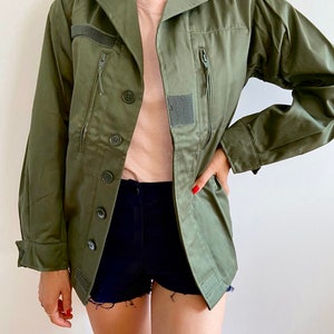 May include: An olive green utility jacket featuring a collared neckline, long sleeves with buttoned cuffs, and a front button closure. The jacket includes two vertical zippered chest pockets, shoulder epaulets, and small rectangular velcro patches.