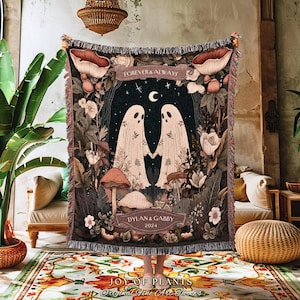 May include: A woven throw blanket featuring a whimsical design with two ghost figures holding hands under a crescent moon and stars. The blanket has a dark background with floral and mushroom accents, and the text "FOREVER & ALWAYS" and "DYLAN & GABBY 2024".