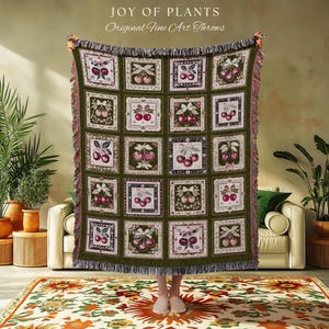 May include: A woven throw blanket featuring a grid of square panels, each displaying a different illustration of cherries with bows. The blanket has a dark green border and a fringed edge. Text at the top reads JOY OF PLANTS Original Fine Art Throws.