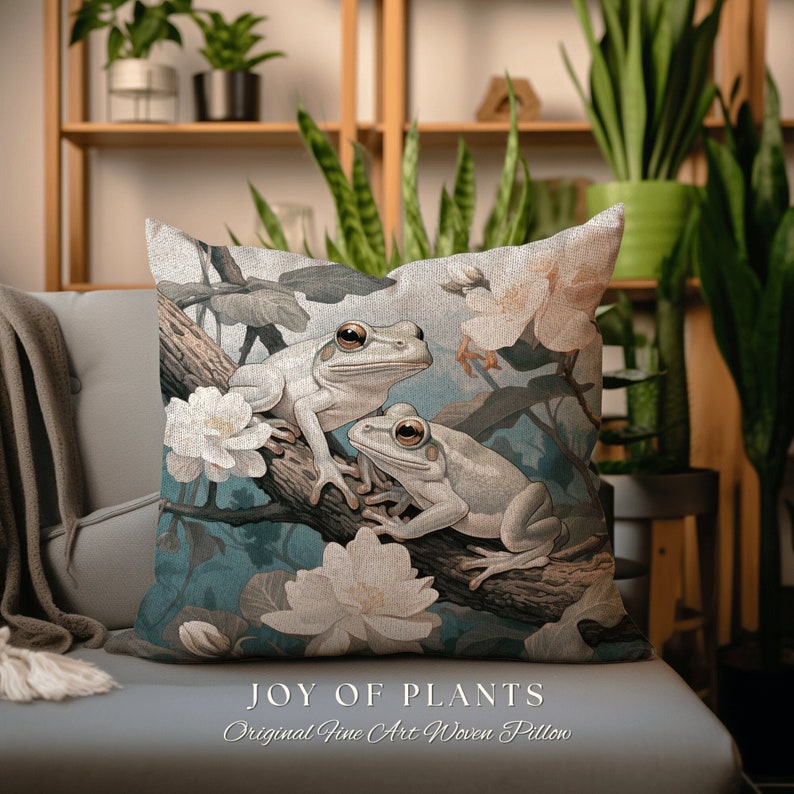May include: A decorative pillow featuring a woven design of two white tree frogs perched on a branch with white flowers. The pillow has a blue background and the text "JOY OF PLANTS Original Fine Art Woven Pillow" is printed at the bottom.