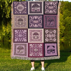 Puede incluir: Una manta tejida con un patrón repetido de cuadrados con setas y calabazas en tonos morados, lavanda y crema. La manta tiene un borde con flecos y el texto "JOY OF PLANTS Original Fine Art Throws" en la parte inferior.