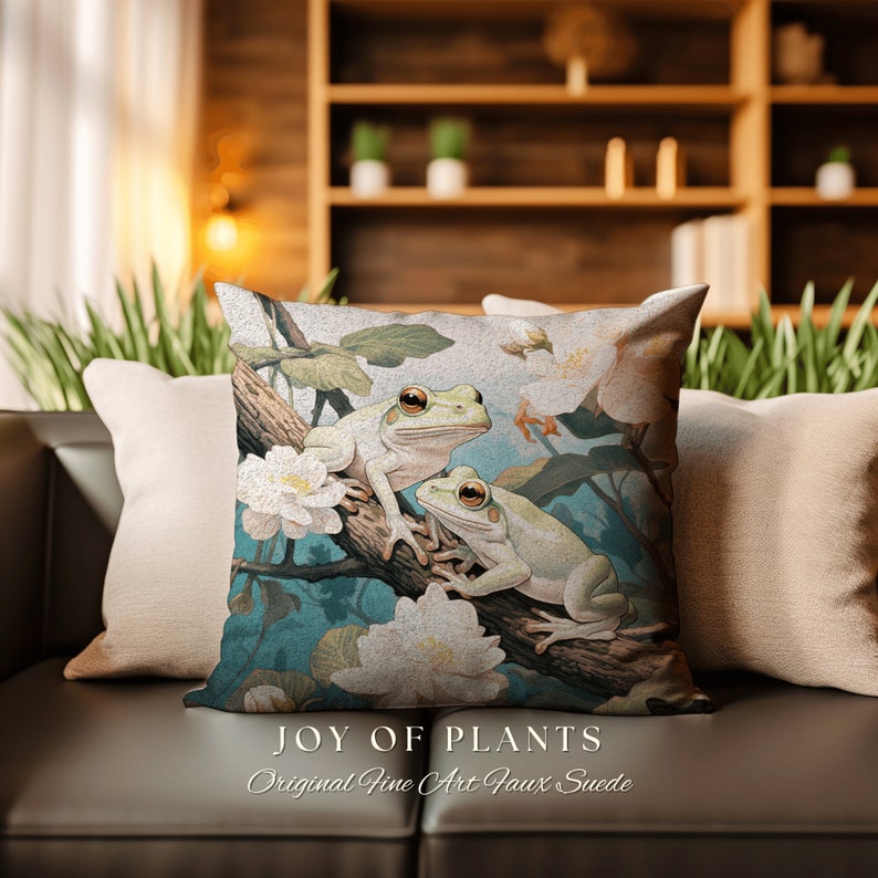 May include: A throw pillow with a white background featuring two green tree frogs sitting on a branch with white flowers. The pillow has the text "Joy of Plants" and "Original Fine Art Faux Suede" printed on the bottom.