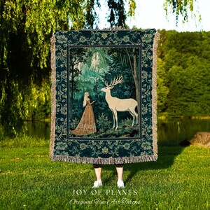 Op de afbeelding: Een tapijtdeken met een bosscène, een vrouw, een hert en bergen. De deken heeft een donkergroene achtergrond met een bloemenrand en franjes. De tekst "JOY OF PLANTS Original Fine Art Throws" staat onderaan.