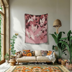 Puede incluir: Una manta rosa con un diseño de murciélagos y uvas colgada en una pared. La manta tiene un borde con flecos y se exhibe sobre un sofá beige con cojines decorativos. El texto "JOPY OF PLANTS Original Fine Art Throws" está en la parte inferior.