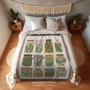 May include: A cream-coloured throw blanket with illustrations of pickle jars. The jars contain pickles and have different lid styles. The blanket has a fringed edge and the text "JOY OF PLANTS Original Fine Art Throws".