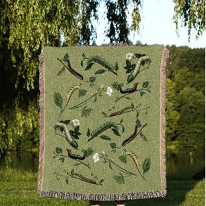 May include: Green throw blanket with a repeating pattern of caterpillars, leaves, and white flowers. The blanket has a fringed edge and the text "JOY OF PLANTS Original Fine Art Throws" at the bottom.
