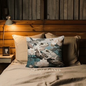 May include: A decorative pillow with a woven design featuring two white tree frogs on a branch with white flowers. The pillow has a blue background and the text "JOY OF PLANTS Original Fine Art Woven Pillow" is printed on the bottom.