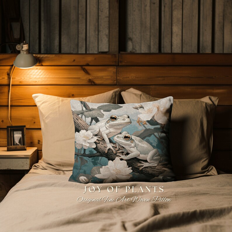 May include: A decorative pillow with a woven design featuring two white tree frogs on a branch with white flowers. The pillow has a blue background and the text "JOY OF PLANTS Original Fine Art Woven Pillow" is printed on the bottom.