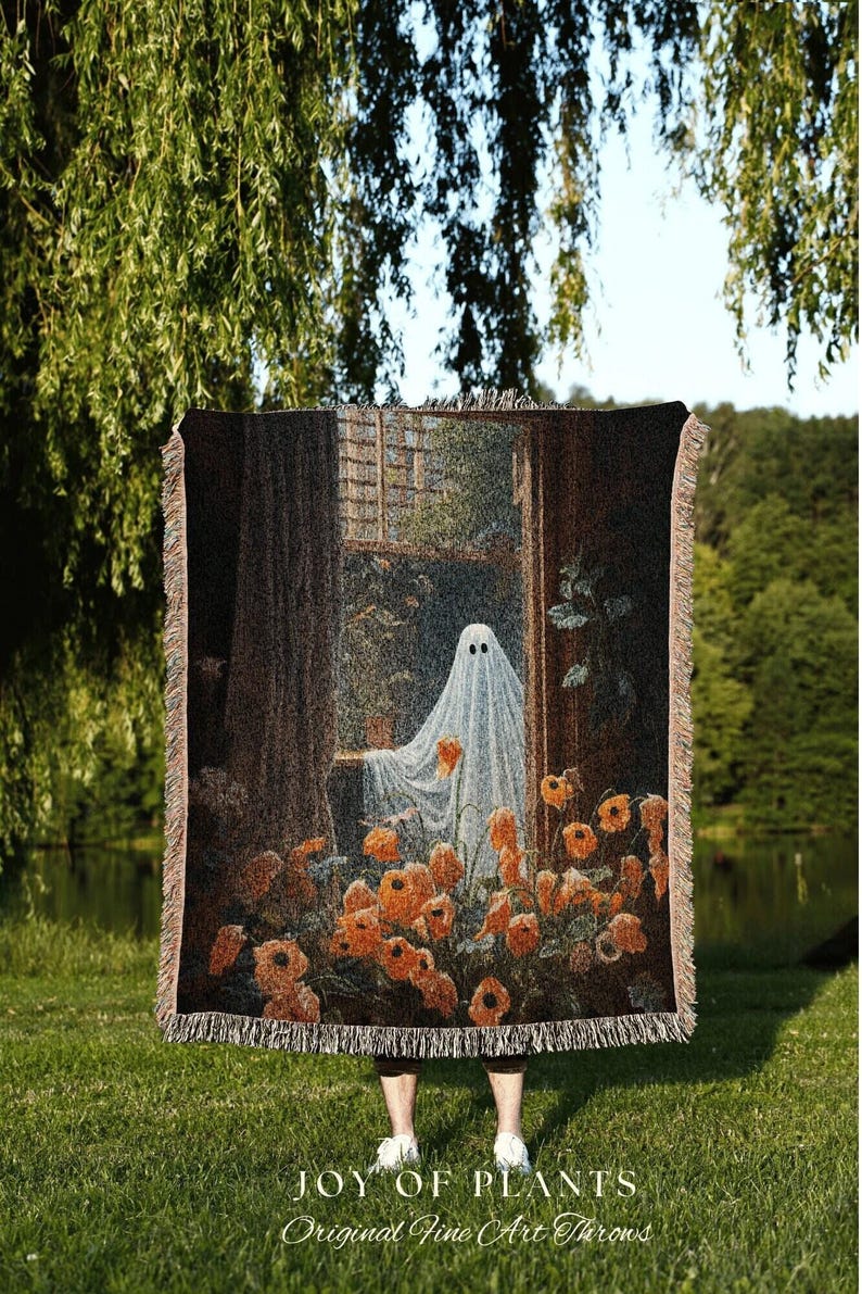May include: A woven throw blanket featuring a whimsical ghost scene with orange flowers and a window. The blanket has a fringed edge and the text "JOY OF PLANTS Original Fine Art Throws" at the bottom. The color palette includes shades of brown, orange, and white.