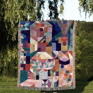 May include: A colorful throw blanket with a geometric design in shades of red, blue, purple, and green. The blanket has a fringed edge and the text "JOY OF PLANTS Original Fine Art Throws" at the bottom.