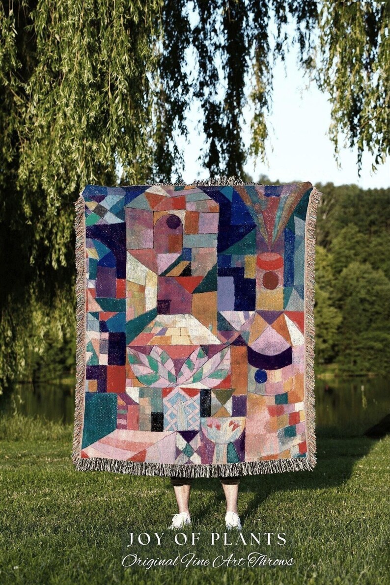 May include: A colorful throw blanket with a geometric design in shades of red, blue, purple, and green. The blanket has a fringed edge and the text "JOY OF PLANTS Original Fine Art Throws" at the bottom.