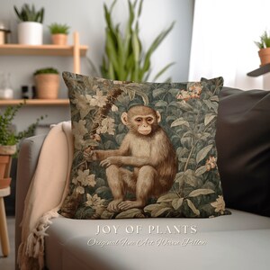 Pode incluir: Uma almofada decorativa com um design de tape&ccedil;aria tecido que representa um macaco sentado em um ambiente exuberante de selva verde. A almofada tem um fundo marrom com folhas verdes e flores rosas. O texto "JOY OF PLANTS" e "Original Fine Art Woven Pillow" est&aacute; impresso abaixo da imagem.