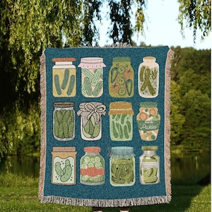 May include: A teal throw blanket with illustrations of pickle jars. The jars contain pickles and other ingredients. The blanket has a fringed edge and the text "JOY OF PLANTS Original Fine Art Throws" at the bottom.