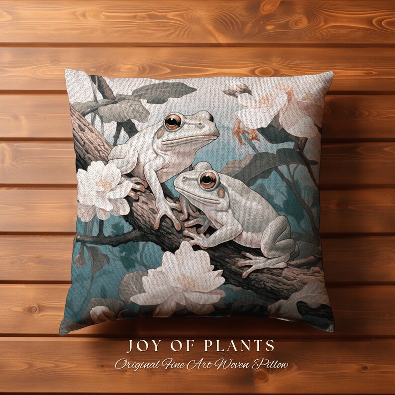 May include: A decorative pillow featuring a woven design of two white frogs sitting on a branch with white flowers. The pillow has a teal background and the text "JOY OF PLANTS Original Fine Art Woven Pillow" at the bottom.