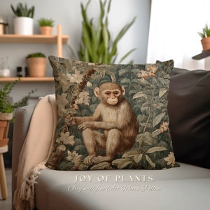 Pode incluir: Uma almofada decorativa com um design de tape&ccedil;aria tecido que representa um macaco sentado em um ambiente exuberante de selva verde. A almofada tem um fundo marrom com folhas verdes e flores rosas. O texto "JOY OF PLANTS" e "Original Fine Art Woven Pillow" est&aacute; impresso abaixo da imagem.