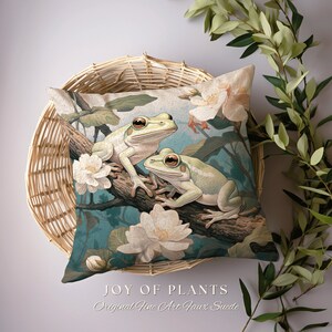 May include: A decorative pillow featuring a green and white illustration of two frogs sitting on a branch with white flowers. The pillow has a light beige background and the text "JOY OF PLANTS Original Fine Art Faux Suede" is printed at the bottom.