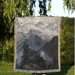 May include: A woven throw blanket featuring a mountain landscape scene in shades of gray, blue, and black. The blanket has a fringed edge and the text "JOY OF PLANTS Original Fine Art Throws" at the bottom.
