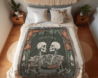 May include: A cream-colored throw blanket with a green and orange design featuring two skeletons embracing, with the text "Eternally Yours" and "Janet & Kenneth". The blanket is on a bed with white pillows and a macrame wall hanging.