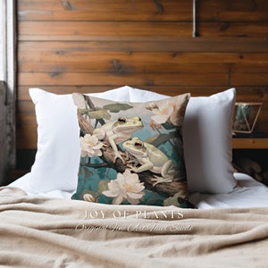 May include: A decorative pillow with a green and white frog design. The frogs are sitting on a branch with white flowers. The pillow is made of faux suede and has the text "JOY OF PLANTS Original Fine Art Faux Suede" printed on the bottom.
