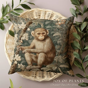 Pode incluir: Uma almofada quadrada com um design tecido que apresenta um macaco sentado em um ambiente exuberante de selva verde. O macaco &eacute; marrom com um rosto marrom claro e est&aacute; cercado por folhas verdes e flores brancas. A almofada tem o texto "JOY OF PLANTS Original Fine Art Woven Pillow" na parte inferior.