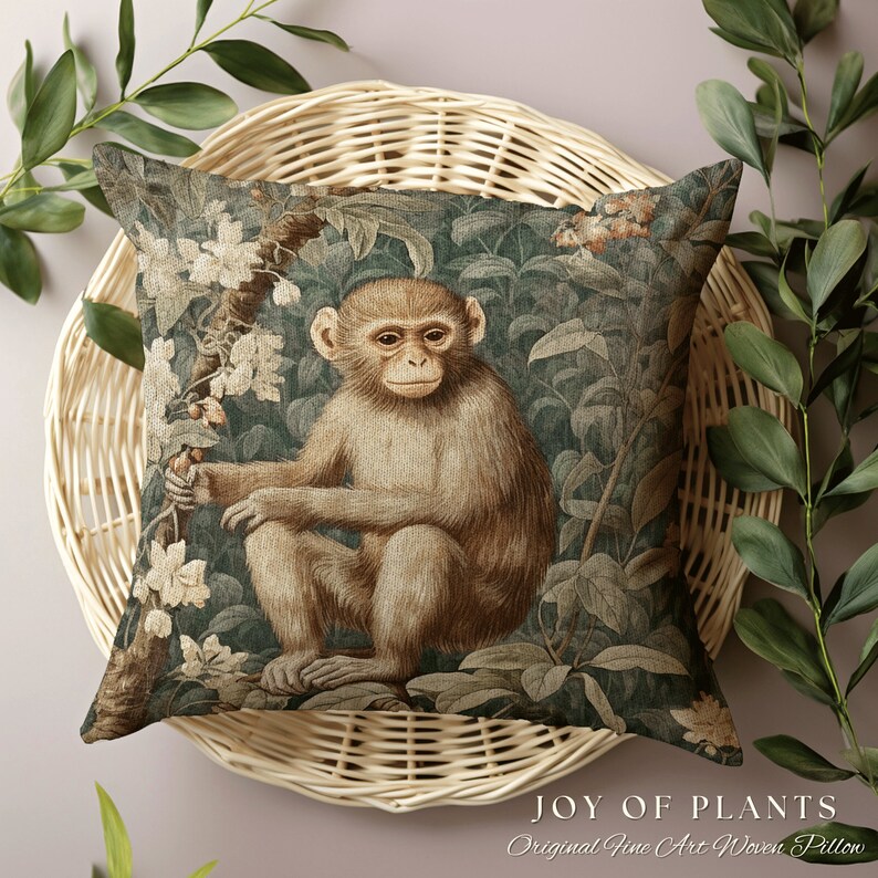 Pode incluir: Uma almofada quadrada com um design tecido que apresenta um macaco sentado em um ambiente exuberante de selva verde. O macaco &eacute; marrom com um rosto marrom claro e est&aacute; cercado por folhas verdes e flores brancas. A almofada tem o texto "JOY OF PLANTS Original Fine Art Woven Pillow" na parte inferior.