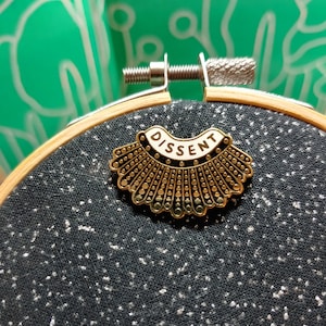 May include: A gold-colored pin in the shape of a judicial collar, with the word "DISSENT" in white letters. The pin is set on a black fabric speckled with white flecks, held in a wooden embroidery hoop. The background is a green fabric with a white pattern.
