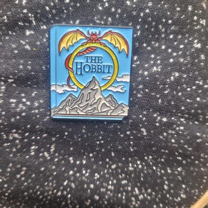 May include: A blue enamel pin featuring the cover of "The Hobbit" book. The pin has a dragon with yellow wings above the title, a mountain range, and white clouds. The pin is set against a black speckled background.
