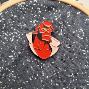 May include: Red and black enamel pin featuring a cartoon character with a red head and a yellow eye. The character is wearing a white shirt and has a red body. The pin is on a black fabric background.