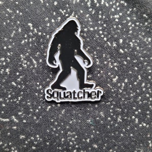 May include: A black and white enamel pin featuring a silhouette of Bigfoot walking, with the word "Squatcher" written below.