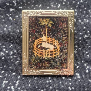 May include: A gold-toned rectangular pin featuring a framed image of a white unicorn inside a wooden fence, with a tree and colorful foliage in the background. The pin has an ornate border and is set against a dark, speckled fabric.