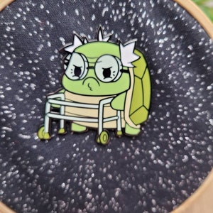 May include: A green turtle enamel pin wearing glasses and using a walker. The turtle has a light green shell and is set against a black fabric background with white speckles. The pin is attached to an embroidery hoop.