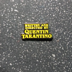 May include: A yellow and black enamel pin with the text "Written and Directed by Quentin Tarantino".